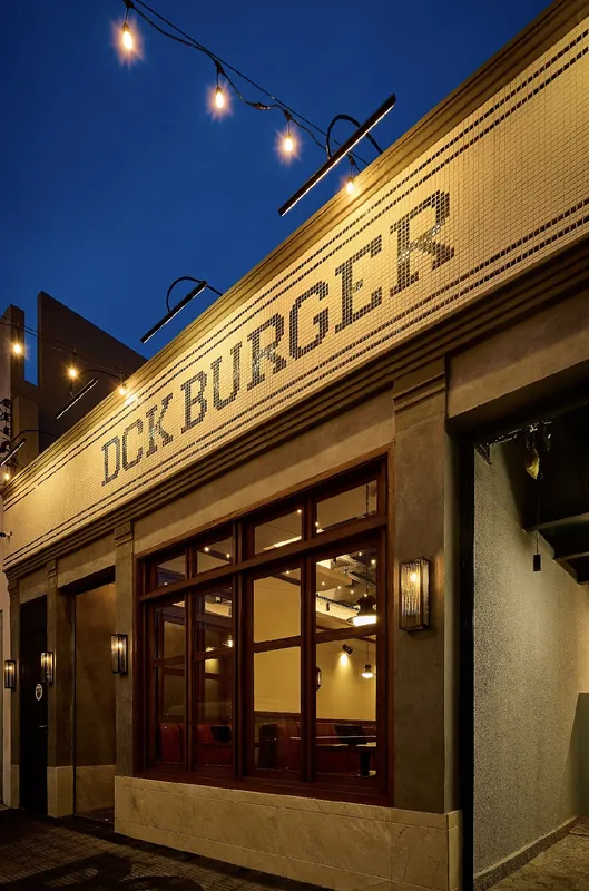 Fachada da DCK Burger ao entardecer, com letreiro em mosaico e bancos vermelhos visíveis nas janelas.