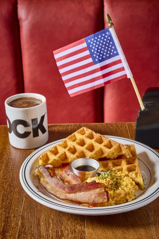 Prato de waffle, bacon e ovos mexidos na DCK, com caneca e bandeira americana sobre a mesa de madeira.