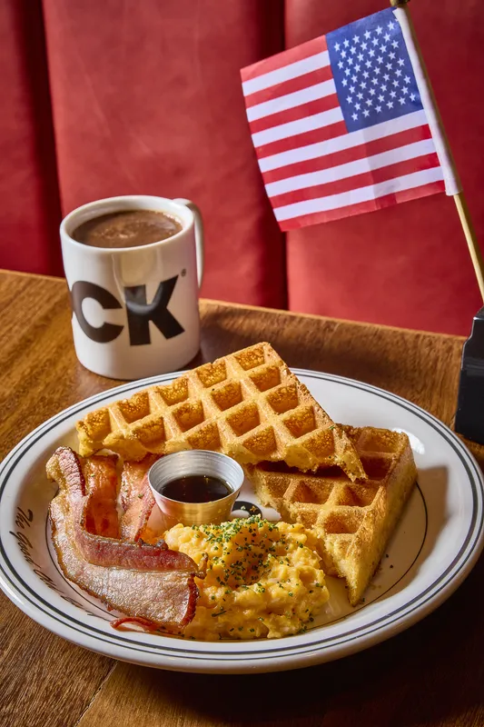 Mesa da DCK vista de cima: waffle, bacon, ovos, xarope, caneca e a cabine vermelha ao fundo.
